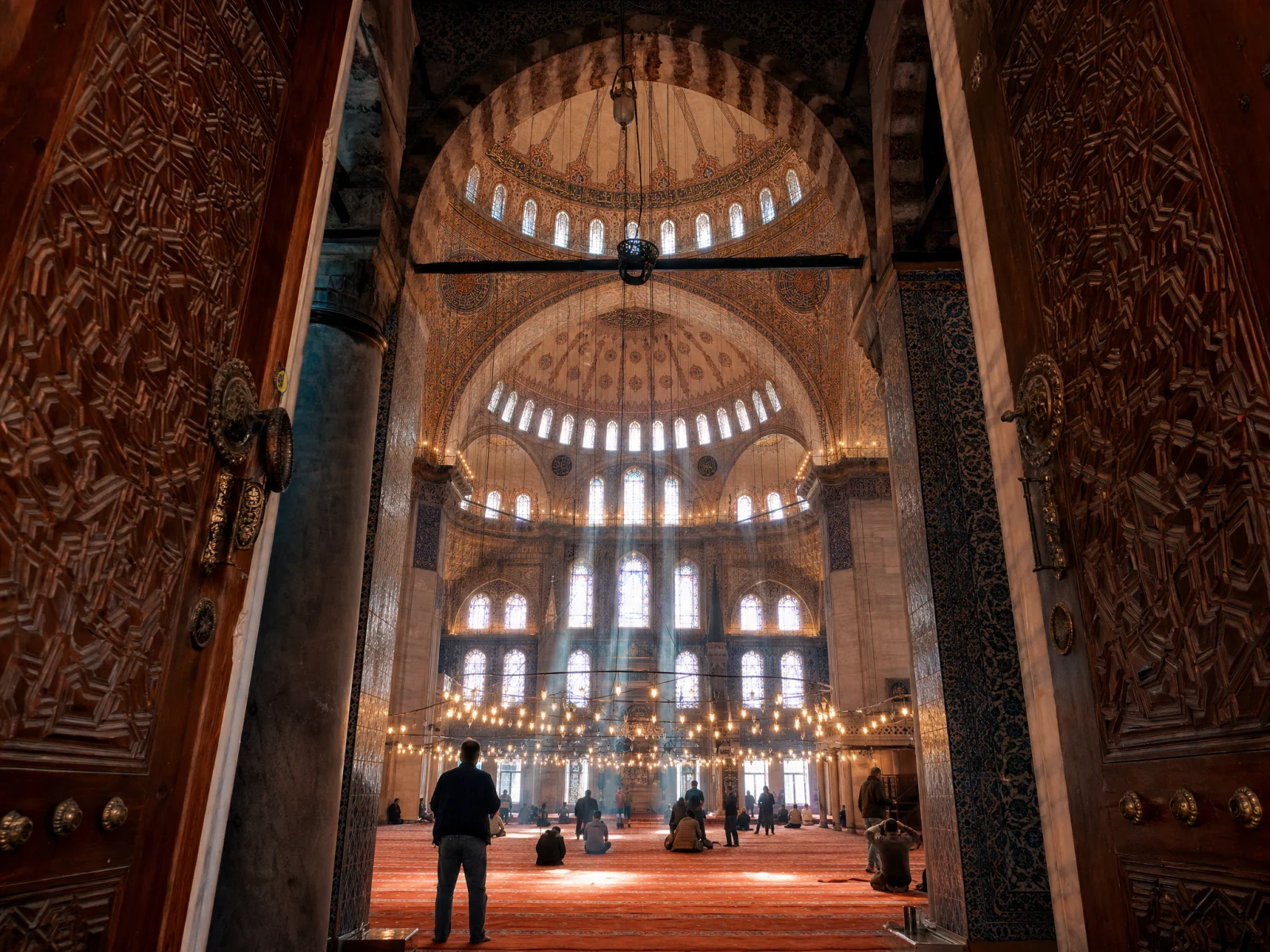 Sunlight streaming through high windows of an Ottoman mosque interior, with intricate domework and pendant lamps overhead